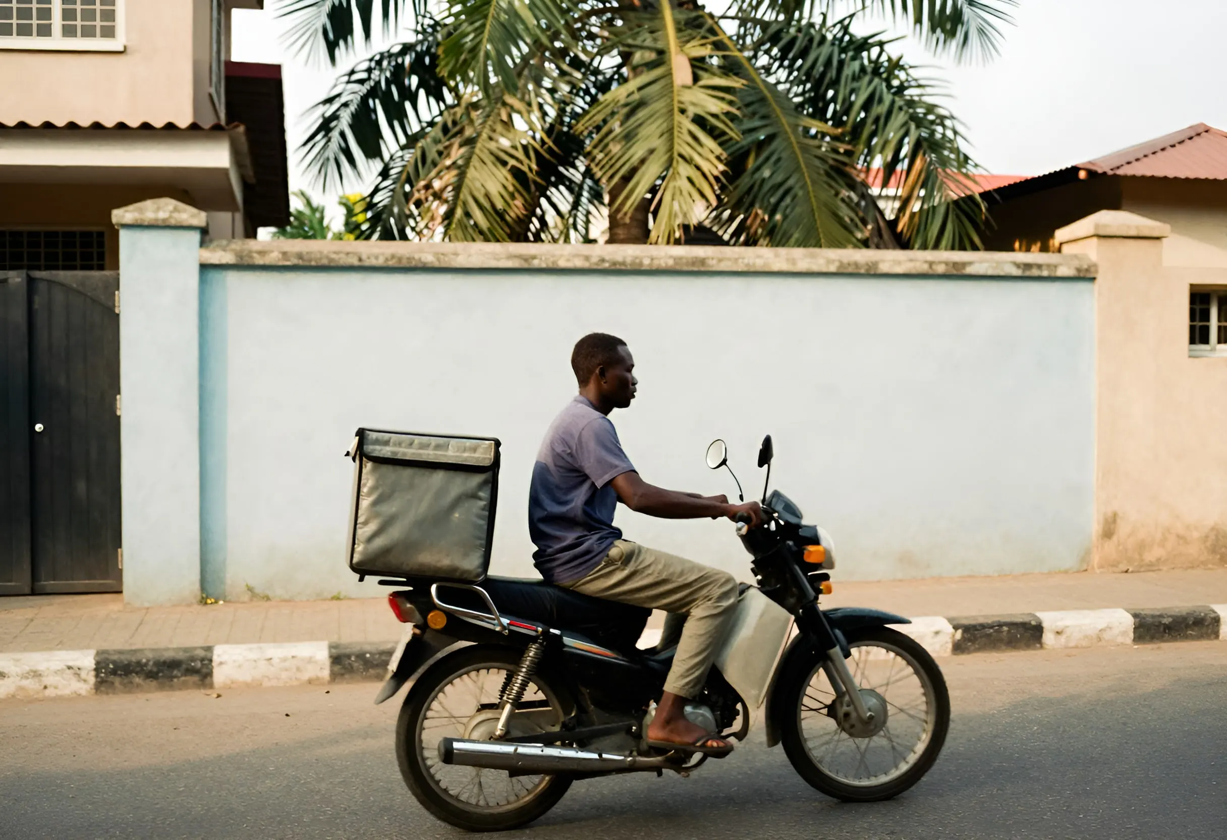 A dispatch rider on a motorcycle with a delivery box, riding past a compound wall